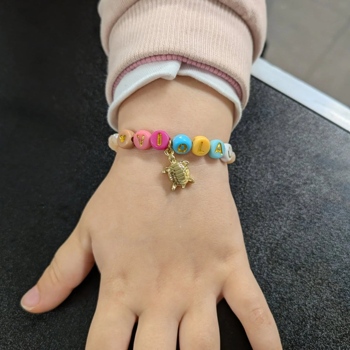 Un bracciale personalizzato con perline-lettera che compongono un nome e un ciondolo dorato, indossato al polso di una bambina.