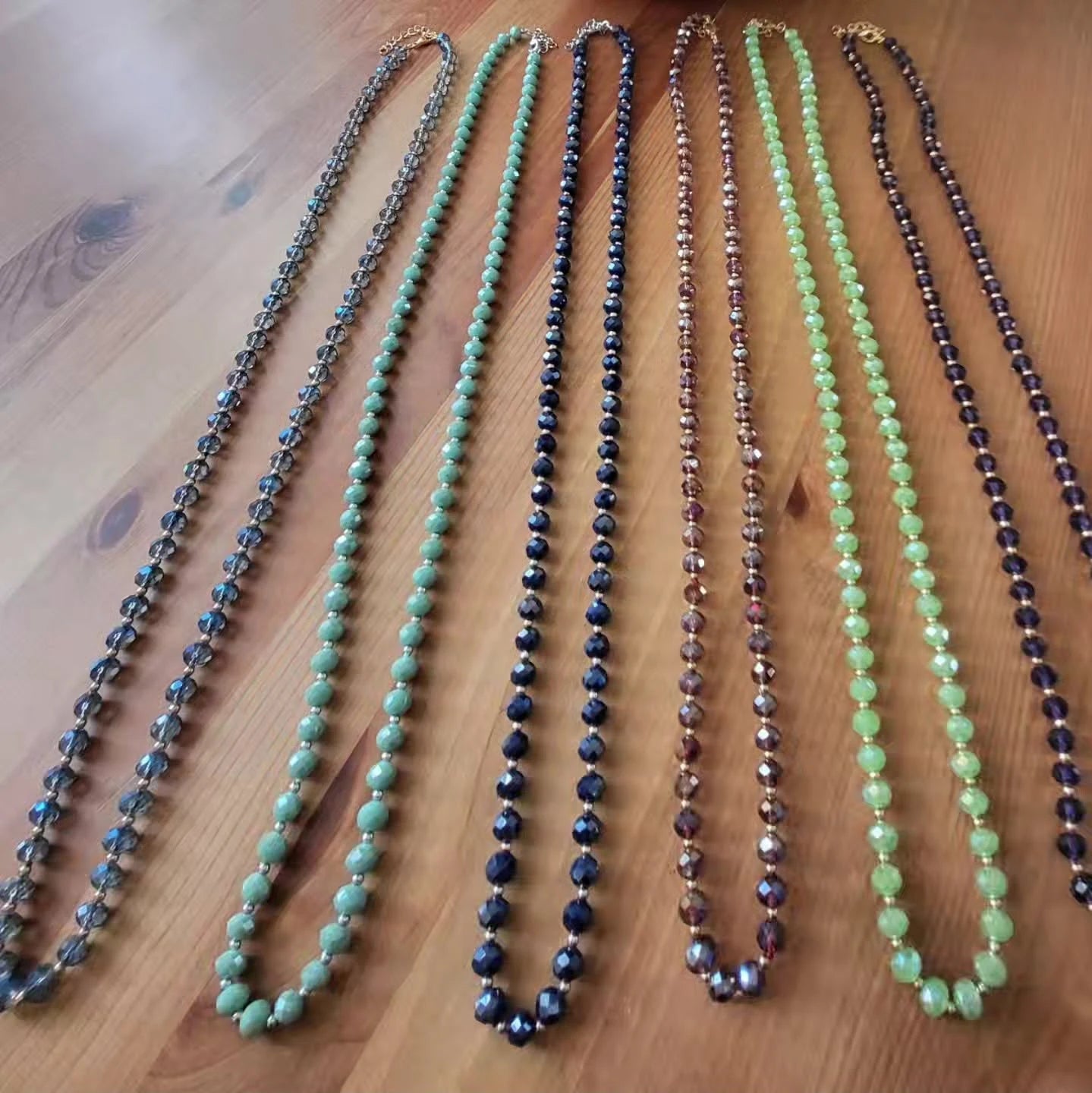 Cinque collane lunghe stese su un piano di legno, ognuna con pietre di un colore diverso: verde giada, grigio ematite, blu notte.