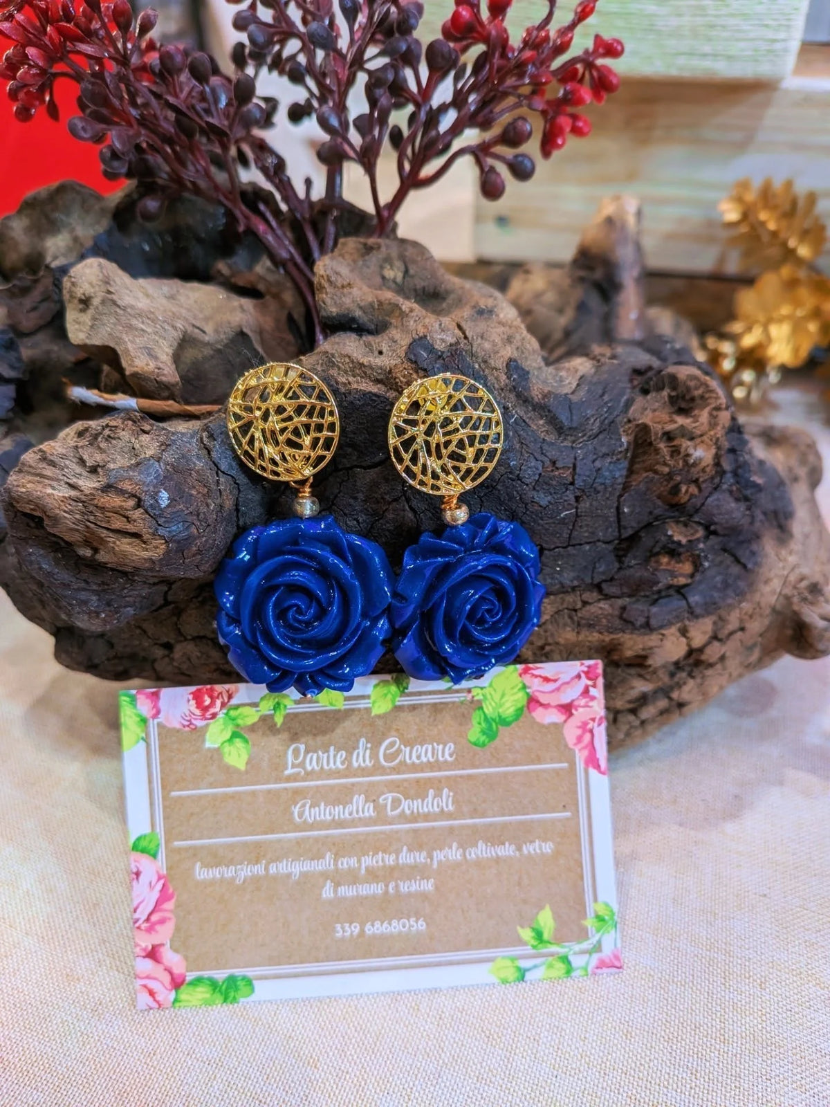 Un paio di orecchini con rosa in resina blu e perno dorato traforato, appoggiati su un ceppo di legno accanto al cartellino della ditta.