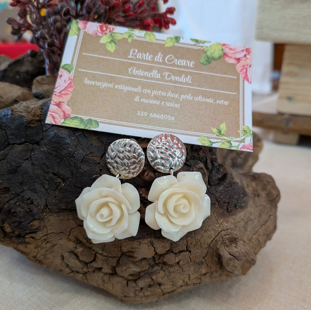 Orecchini con rosa in resina bianco avorio e perno argentato inciso, su un ceppo di legno con il cartellino di L'arte di creare.