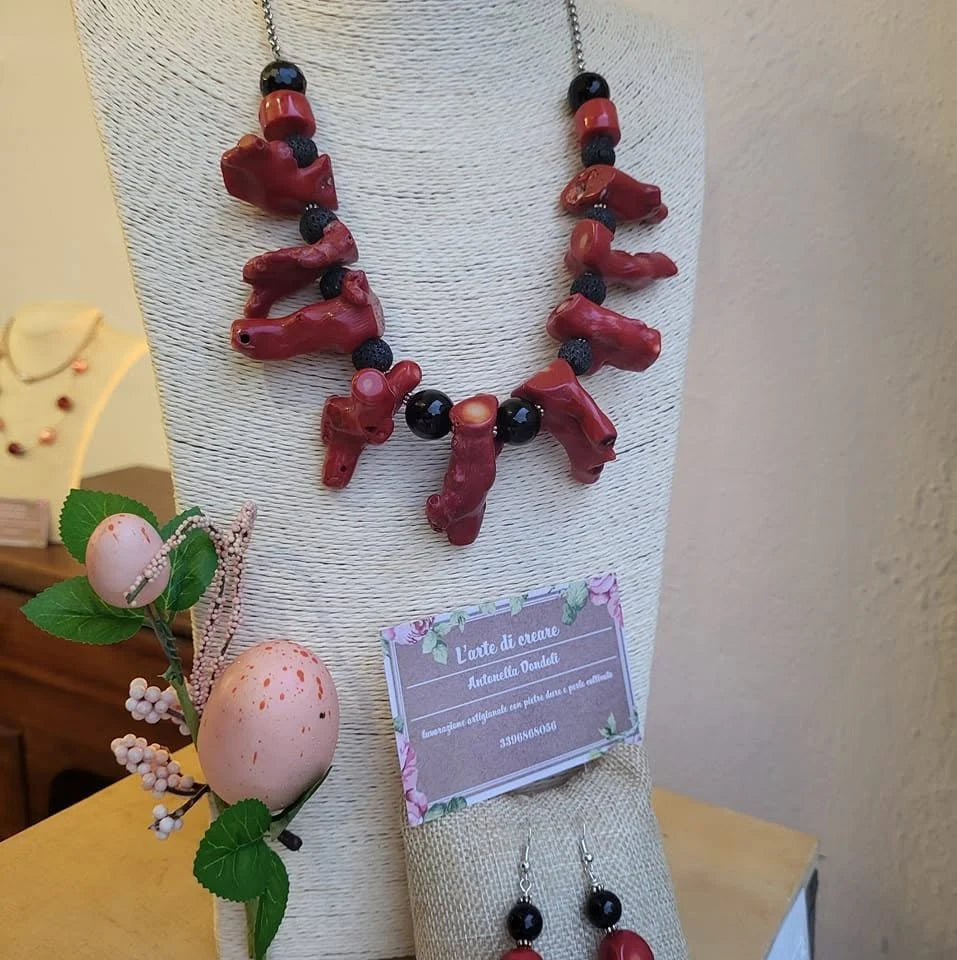 Una parure di collana e orecchini in corallo rosso e pietre nere su espositore di juta, accanto al cartellino di L'arte di creare.