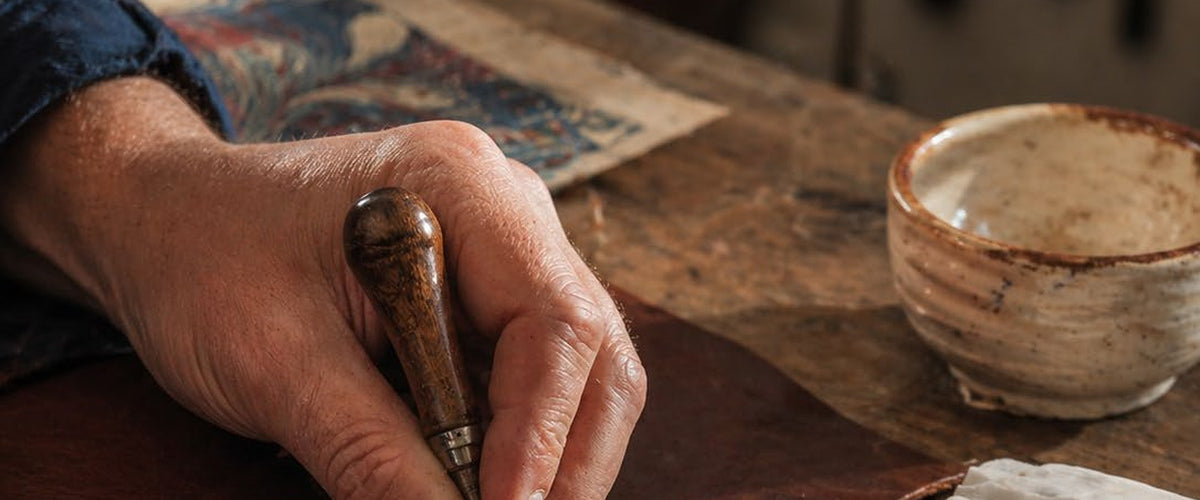 La mano di un artigiano incide il cuoio con una lesina, su un banco di legno consumato